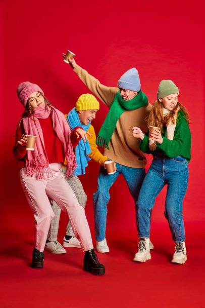 Groep jongeren, vrienden in winterkleding die samen plezier hebben, geïsoleerd poseren over rode achtergrond. Dansen. Concept van emoties, wintervakantie, mode, levensstijl, feest, vriendschap - Foto, afbeelding