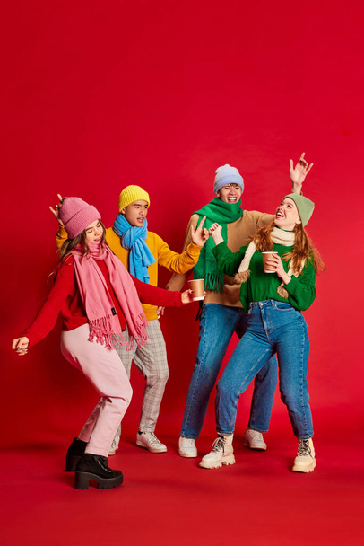 Jongeren, vrienden in winterkleding die samen plezier hebben, geïsoleerd poseren over rode achtergrond. Vrolijke feestdagen. Concept van emoties, wintervakantie, mode, levensstijl, feest, vriendschap - Foto, afbeelding