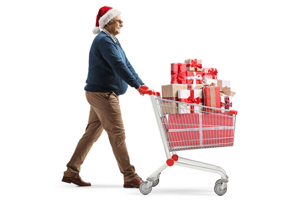 Full length profile shot of a mature man with a santa hat pushing a shopping cart with presents isolated on white background - Φωτογραφία, εικόνα