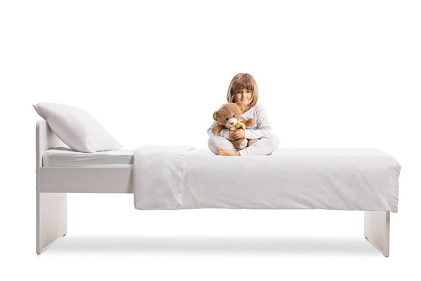 Little girl in pajamas hugging a teddy bear and sitting on a bed isolated on white background - Photo, image