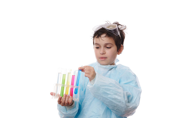 Κοντινό πλάνο ενός πολύ-εθνοτικού έξυπνου έφηβου αγοριού, ενός νεαρού χημικού φοιτητή που φοράει μπλε εργαστηριακή ποδιά και γυαλιά, με δοκιμαστικούς σωλήνες σε τρίποδο, που διεξάγει πειράματα χημείας, απομονωμένο σε λευκό φόντο - Φωτογραφία, εικόνα