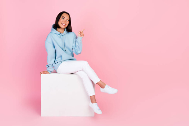Full length photo of cute dreamy lady wear blue hoodie sitting white platform pointing empty space isolated pink color background. - Φωτογραφία, εικόνα
