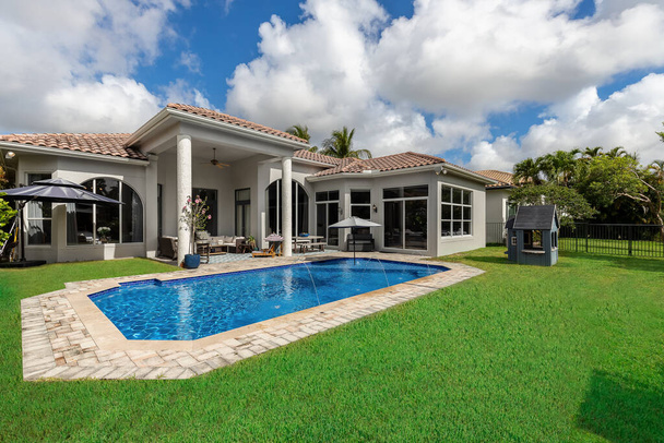 Dvůr elegantního domu s bazénem, krátká tráva, venkovní nábytek, deštník, dům s šedými stěnami a bílými detaily, střecha s červenými dlaždicemi, modrá obloha s mraky - Fotografie, Obrázek