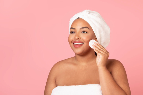 Skincare routine. Happy black chubby woman cleansing skin with cotton pad, beautiful body positive lady removing makeup from face, standing wrapped in towel on pink background - Photo, Image