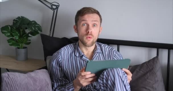 Emoción conmocionada, sorprendida o confusa: atractivo hombre barbudo usando tablet computer sentado en la cama por la mañana. Hombre caucásico en ropa de dormir usando touchpad. Video de alta calidad 4k - Metraje, vídeo
