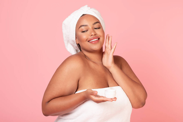 Skin care concept. Happy black oversize lady with towel on head applying moisturizing face cream, beautiful african american obese woman standing over pink background - Фото, зображення