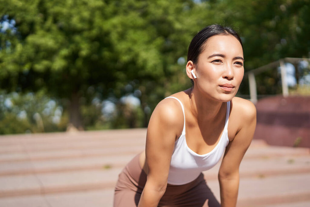 Portret Azjatki podczas przerwy, ciężko oddychającej i dyszącej po bieganiu, biegającej i wycierającej pot z czoła, uśmiechniętej zadowolonej. - Zdjęcie, obraz