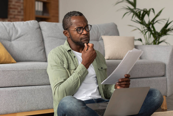 集中型の中年アフリカ系アメリカ人男性で、ノートパソコン付きのメガネにはリビングルームのインテリアに文書が読み込まれます。仕事とビジネス、新しい通常、会計とデータ分析、スタートアップの帳簿作成 - 写真・画像