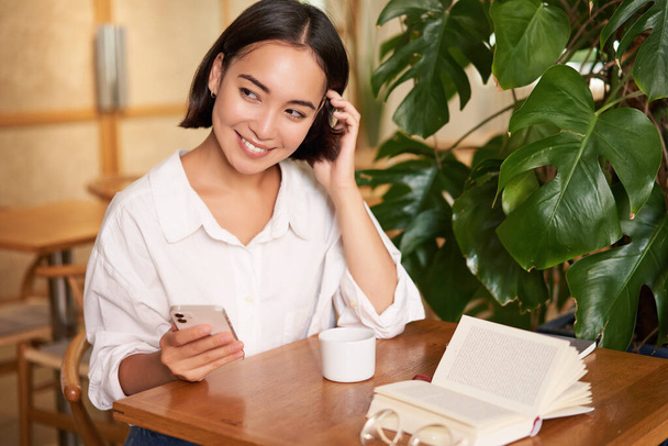 Piękna młoda kobieta w kawiarni z filiżanką kawy i książki, trzymając smartfona, za pomocą telefonu komórkowego, płacąc bezstykowe czekanie na kelnera. - Zdjęcie, obraz