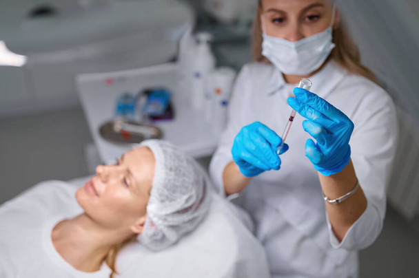 Injection cosmetology at professional beauty salon concept. Focus on cosmetologist hand in sterile glove holding syringe and flask with vitamin - Photo, Image