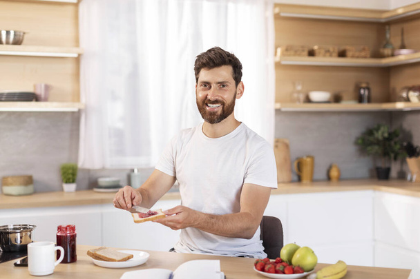 Vidám, középkorú fehér férfi szakállal fehér pólóban szendvicset készít gyümölcsökkel reggelire, élvezi a hétvégét, szabadidejét a modern konyha belsejében. Jó reggelt egyedül, egészséges étel otthon. - Fotó, kép