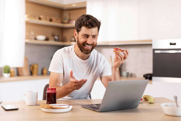Smiling middle aged caucasian male with beard in white t-shirt eats sandwich, has video call, gesticulates in kitchen interior. Good morning and breakfast, meeting remotely, social distance at home - Foto, afbeelding