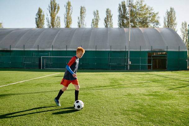 Elementary age football forward kicking ball. Junior boy soccer player training along on field before competition match at school sport club - Foto, afbeelding