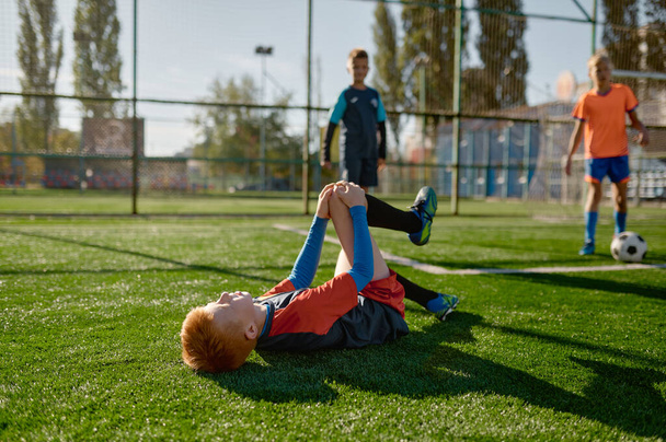 Young boy soccer player with injured knee lying on field during match. Hurt kid footballer with painful leg on ground in agony having bad day on pitch - Fotografie, Obrázek