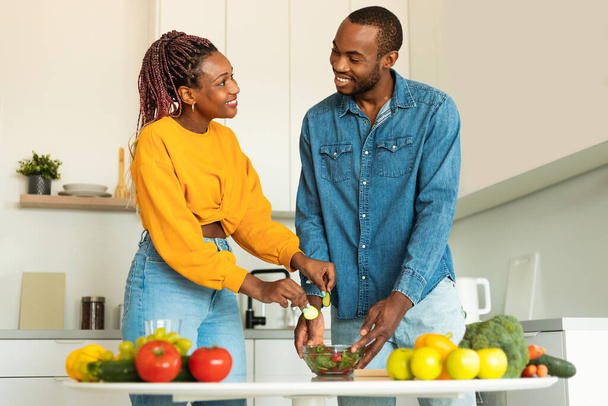 Αγαπώντας τους Αφροαμερικανούς συζύγους που μαγειρεύουν μαζί στη σύγχρονη κουζίνα στο σπίτι, φτιάχνοντας υγιεινή σαλάτα για δείπνο, κοιτώντας και χαμογελώντας ο ένας στον άλλο. Διατροφή και οικογενειακές συνταγές - Φωτογραφία, εικόνα