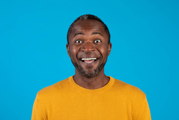Closeup portrait of excited middle aged handsome black man in casual cheerfully smiling with eyes and mouth wide open over blue studio background, copy space. Happiness, excitement, human emotions - Photo, Image