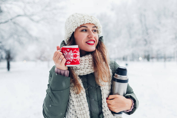 Portrait of young happy woman drinking hot tea holding vacuum flask in snowy winter park enjoying landscape under falling snow. Cup dressed in red knitted Christmas case - Photo, Image