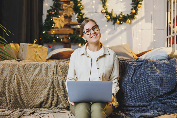 A beautiful woman works at home on New Year's holidays, wear comfortable home clothes, look at a laptop, work remotely for the New Year, plaid, New Year's winter mood. - Foto, imagen