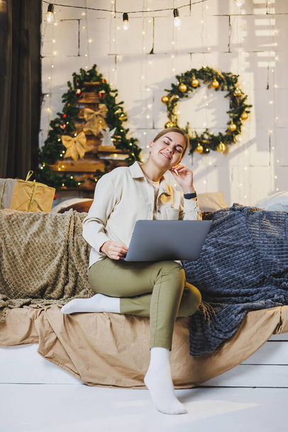 A beautiful woman works at home on New Year's holidays, wear comfortable home clothes, look at a laptop, work remotely for the New Year, plaid, New Year's winter mood. - Photo, Image