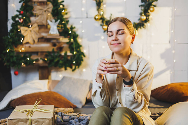 Portret uśmiechniętej kobiety z pachnącą kawą w rękach na tle noworocznych dekoracji. Świąteczna kawa w domu na łóżku, Święta Nowego Roku - Zdjęcie, obraz