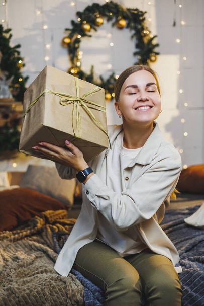 Boldog csinos nő, szép hajjal, kényelmes ruhákban, szilveszteri ajándékkal a kezében az ágyon ül a karácsonyfa mellett. Újévi hangulat otthon. - Fotó, kép
