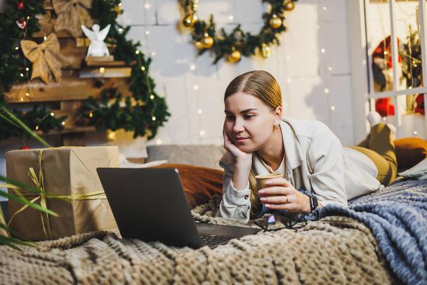 Pretty woman with blond hair in cozy clothes with coffee in hand using laptop working remotely and smiling on bed in room with Christmas tree at home. New Year holidays. - Fotoğraf, Görsel
