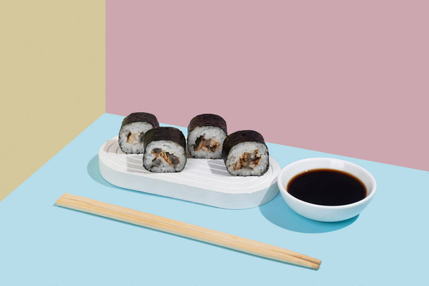 Hosomaki (sushis, petits pains) à l'anguille et sauce soja sur un support en plâtre blanc sur un fond uni coloré (bleu, rose, jaune). Une composition simple et concise - Photo, image