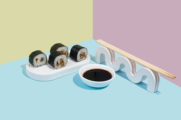 Hosomaki (sushi, rolls) with eel and soy sauce on a white plaster stand on a colorful plain background (blue, pink, yellow). A simple concise composition - Photo, Image