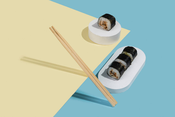 Hosomaki (sushi, rollos) con anguila sobre un soporte de yeso blanco sobre un fondo liso multicolor (azul, amarillo). Una composición simple y concisa - Foto, Imagen