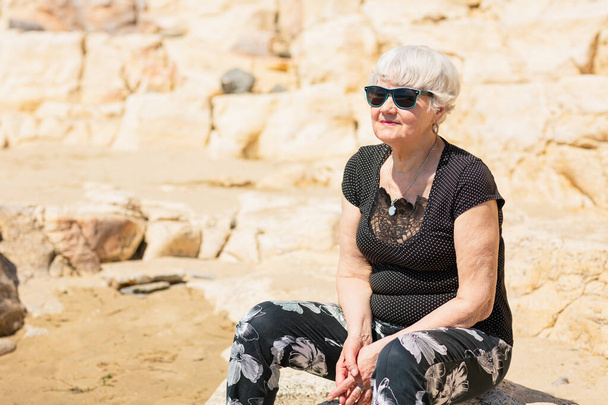 Old woman in black clothes sitting on a rock near the sea and having fun. - Φωτογραφία, εικόνα