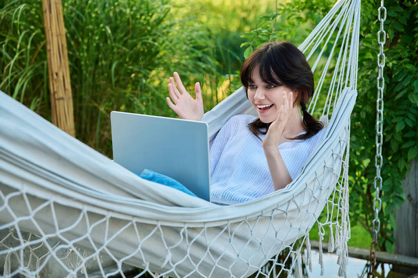 Young teenage female lying outdoor in hammock with laptop talking with friends relatives via video call. Happy girl 18, 19 years old relaxing talking online using webcam at conference - Fotó, kép