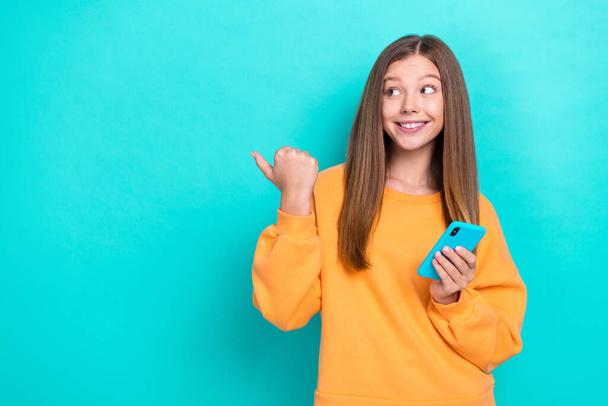 Photo of young schoolgirl wear sweater thumb directing empty space hold phone online shopping black friday isolated on aquamarine color background. - Valokuva, kuva