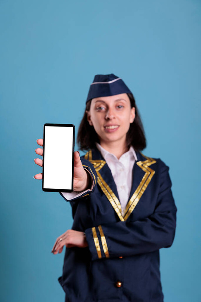 Glimlachende stewardess toont moderne telefoon met blanco wit scherm, smartphone app reclame mockup. Vliegtuiggastvrouw met telefoon met leeg display voor webpromotie - Foto, afbeelding