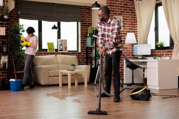 集中した女性は装飾的なオブジェクトをほこりながら幸せなアフリカ系アメリカ人男性が床を真空。異人種間のカップルが調和して家を掃除し、家事を一緒にする. - 写真・画像