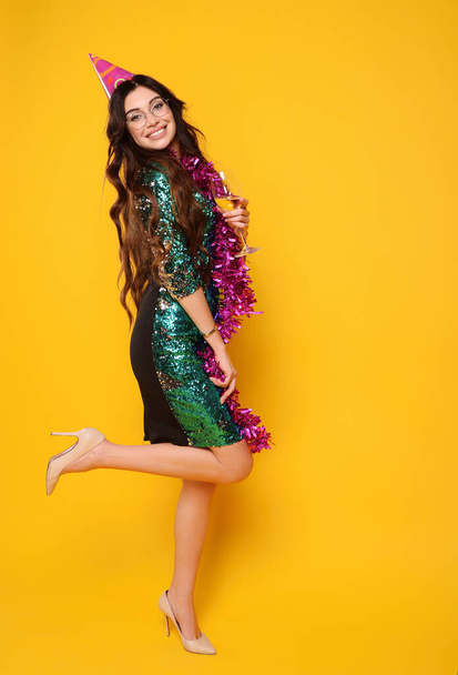 pretty young woman in a smart dress in a party cap with a pink boa around her neck and a glass of champagne smiling on a yellow background - Foto, Bild