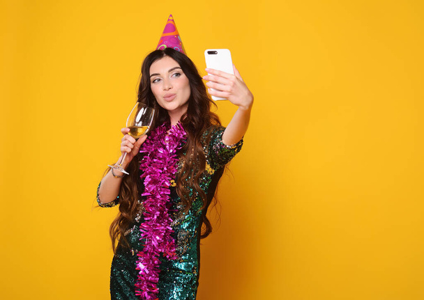 young pretty woman in a pink party hat and pink boa holds a glass of champagne in her hands, smiles and holds a smartphone in her hands - Photo, Image