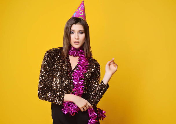 young pretty woman in golden dress wearing pink boa and party cap on yellow background - Foto, immagini