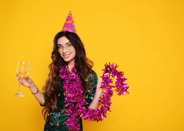 pretty young woman in a smart dress in a party cap with a pink boa around her neck and a glass of champagne smiling on a yellow background - Foto, imagen