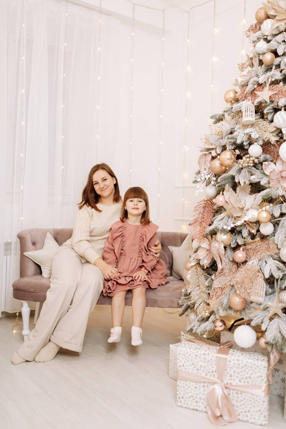 Καλά Χριστούγεννα και καλές γιορτές. Χαρούμενη μαμά και χαριτωμένες κόρες κορίτσι της αγκαλιάζει και χαμογελά στον καναπέ. Γονείς και μικρά παιδιά διασκεδάζουν και παίζουν μαζί κοντά στο χριστουγεννιάτικο δέντρο σε εσωτερικούς χώρους. - Φωτογραφία, εικόνα