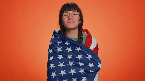 Junge Nerd-Frau mit Zöpfen in Gläsern, die die amerikanische Flagge schwenken und einwickeln, feiern, Menschenrechte und Freiheiten. Unabhängigkeitstag. Latin erwachsene Mädchen isoliert auf Studio orange Wand Hintergrund - Foto, Bild