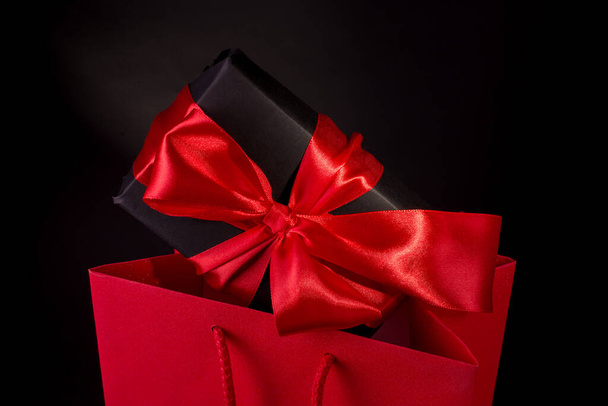 Χαρούμενη Ημέρα του Αγίου Βαλεντίνου ή Black Friday Shopping Concept με μαύρο κουτί δώρου και κόκκινο σατέν τόξο κατά μαύρο φόντο. Διαφημιστικό banner με χώρο αντιγραφής. - Φωτογραφία, εικόνα