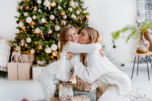 Cute mother with daughter are hugging near christmas gifts next to christmas tree at home - Fotó, kép