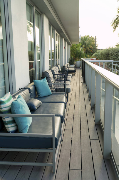 Reihe von Outdoor-Sofas mit Stahlrahmen in der Nähe der reflektierenden Glaswände - Miami, Florida. Stahlsofa auf einem Holzdeck mit bemalten Geländern und Blick nach draußen mit Bäumen. - Foto, Bild