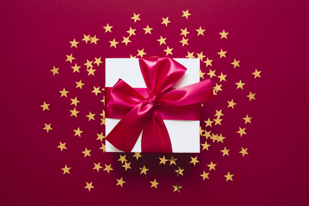 Λευκό κουτί δώρου με κορδέλα σε κόκκινο χρώμα με χρυσά κομφετί σε σχήμα αστεριού. Του Αγίου Βαλεντίνου, Χριστούγεννα ή Πρωτοχρονιά. Επίπεδη lay στυλ με μινιμαλιστικό σχεδιασμό. - Φωτογραφία, εικόνα
