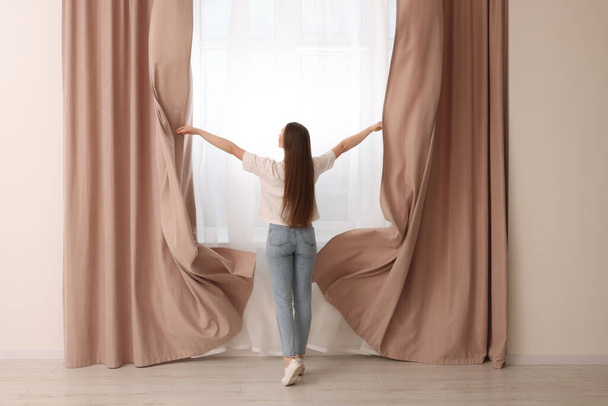 Woman opening stylish curtains at home, back view - Фото, зображення