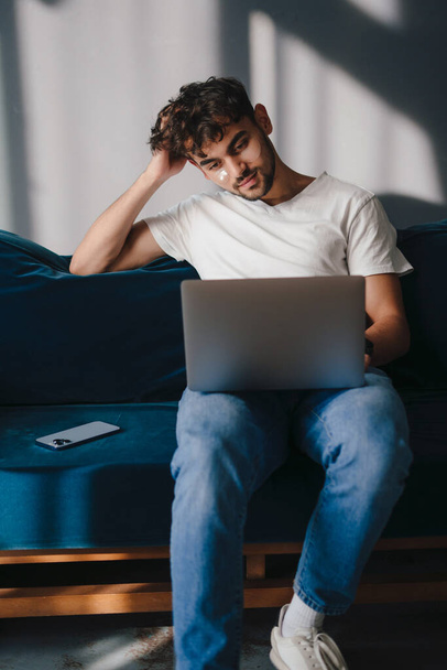 Νεαρός άνδρας που χρησιμοποιεί φορητό υπολογιστή κάθεται σε μπλε καναπέ στο σπίτι ανάπαυσης και χαλάρωσης, ενώ περνούν ελεύθερο χρόνο στο σαλόνι. Τεχνολογική έννοια. Έννοια τρόπου ζωής ανθρώπων - Φωτογραφία, εικόνα