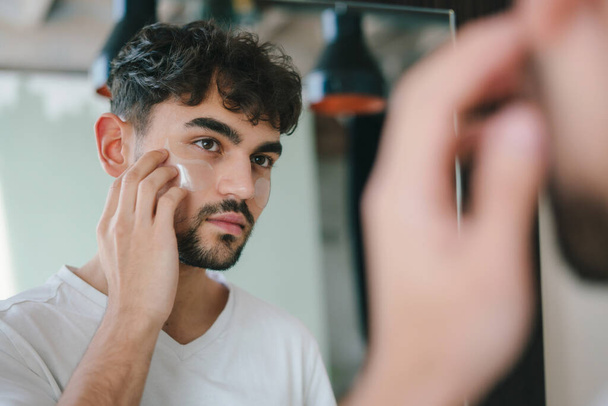 Een man met een baard die oogvlekken onder zijn ogen aanbrengt terwijl hij naar de spiegel kijkt. Rimpels en gezicht thuiszorg voor mannen. Huidverzorging routine. Thuis spa zorg concept. Gezichtsbehandeling - Foto, afbeelding