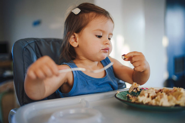 Niña feliz aprendiendo a comerse con un tenedor mientras está sentada en una silla para niños. Sonriente niño come con las manos. - Foto, Imagen