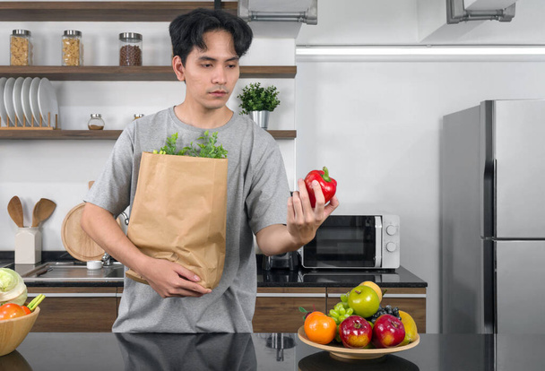 Νεαρός Ασιάτης άνδρας σε casual φορούν κρατώντας το κόκκινο bell pepper ενώ μεταφέρουν χάρτινη τσάντα γεμάτη με λαχανικά. Πρωινή ατμόσφαιρα σε μια σύγχρονη κουζίνα - Φωτογραφία, εικόνα
