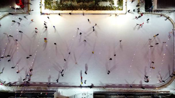 Les gens patinent sur la patinoire à l'extérieur sur la vue de dessus de nuit d'hiver. Vue aérienne sur drone. Lumineuses guirlandes lumineuses lampes éclairage. Jetez de longues ombres. Nouvel An Noël célébration vacances loisirs - Photo, image